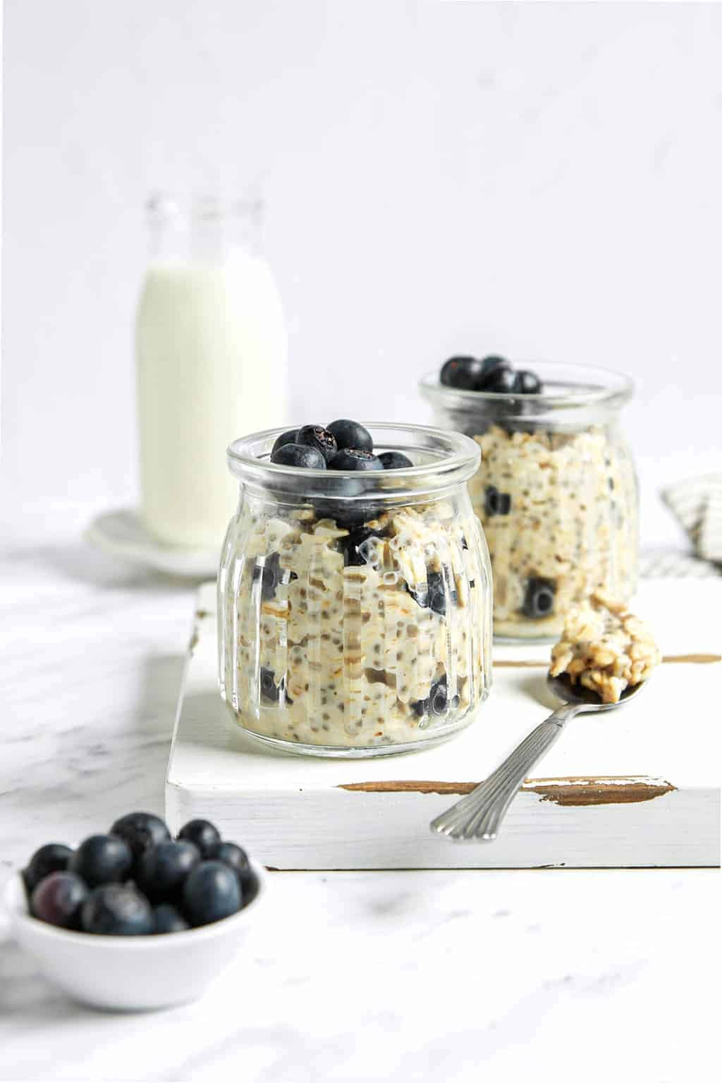 Two jars of overnight oats with blueberries on a white wooden board. 