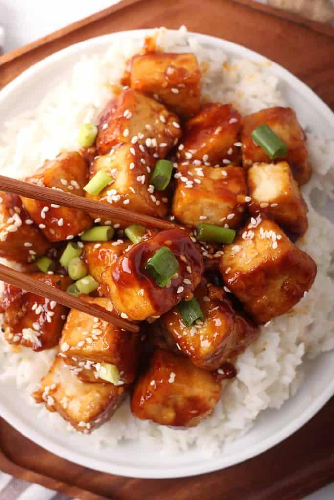 close up shot of general tso's tofu over rice in a white bowl with wooden chopsticks.