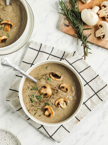 top view of two bowls of vegan dairy-free cream of mushroom soup with whole mushrooms, fresh herbs, salt, pepper, spoon, and checked linens on white surface