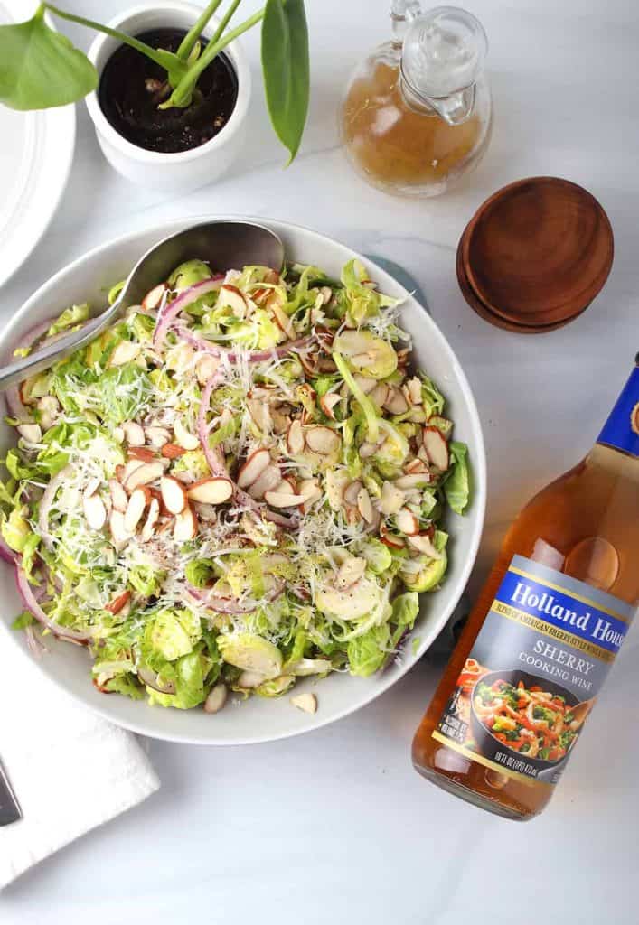overhead shot of white serving bowl filled with shaved brussels sprouts salad topped with sliced almonds and a bottle of holland house sherry cooking wine on a white table