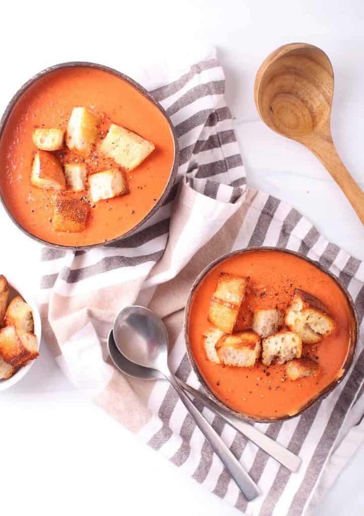 Two bowls of creamy vegan tomato soup with homemade croutons next to two spoons. 