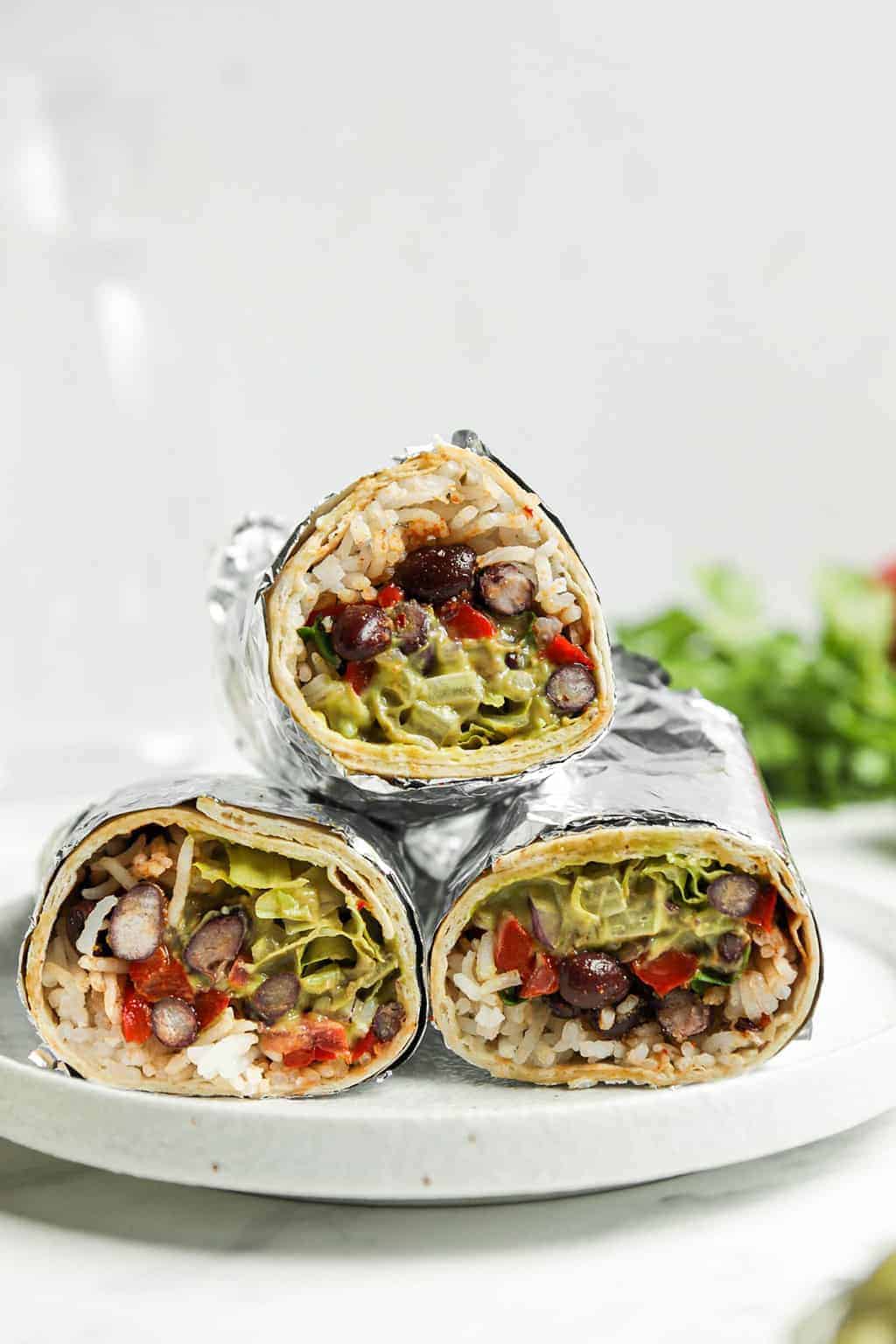 Three vegan burritos wrapped in foil and stacked on a white plate 