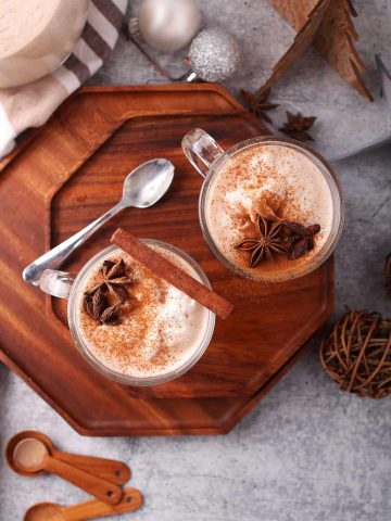 Two glasses of eggnog with whole spices on a wooden platter