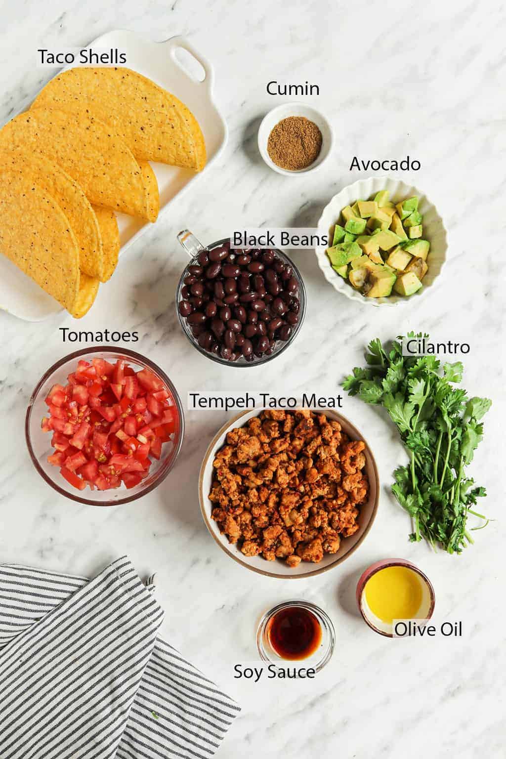 ingredients for homemade tacos including tempeh, cilantro, oil, soy sauce, tomatoes, avocado, black beans, cumin, and taco shells
