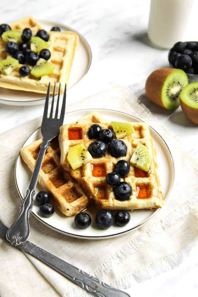 classic vegan waffles served on a white plate with a fork, blueberries, kiwi and maple syrup. 